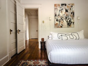 A bedroom with a large bed, a wooden floor, a white door, and a painting on the wall. - Eclectic & Unique Historic Home  (New Albany)