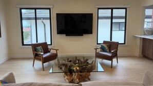 A living room with two leather chairs, a glass coffee table with a wood centerpiece, a mounted TV, and large windows with a view of the street. - Fully Renovated 2 BR House by Palos Verdes, Beach, in Quiet Neighborhood (Los Angeles)
