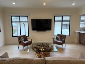 A living room with two leather chairs, a glass coffee table with a wood centerpiece, a mounted TV, and large windows with a view of the street.