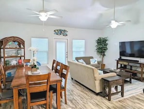 A modern living room with a dining area, featuring a sofa, TV, and ceiling fans. - BEACH~HEATED POOL~PET FRIENDLY-WINTER STAY (Corpus Christi)