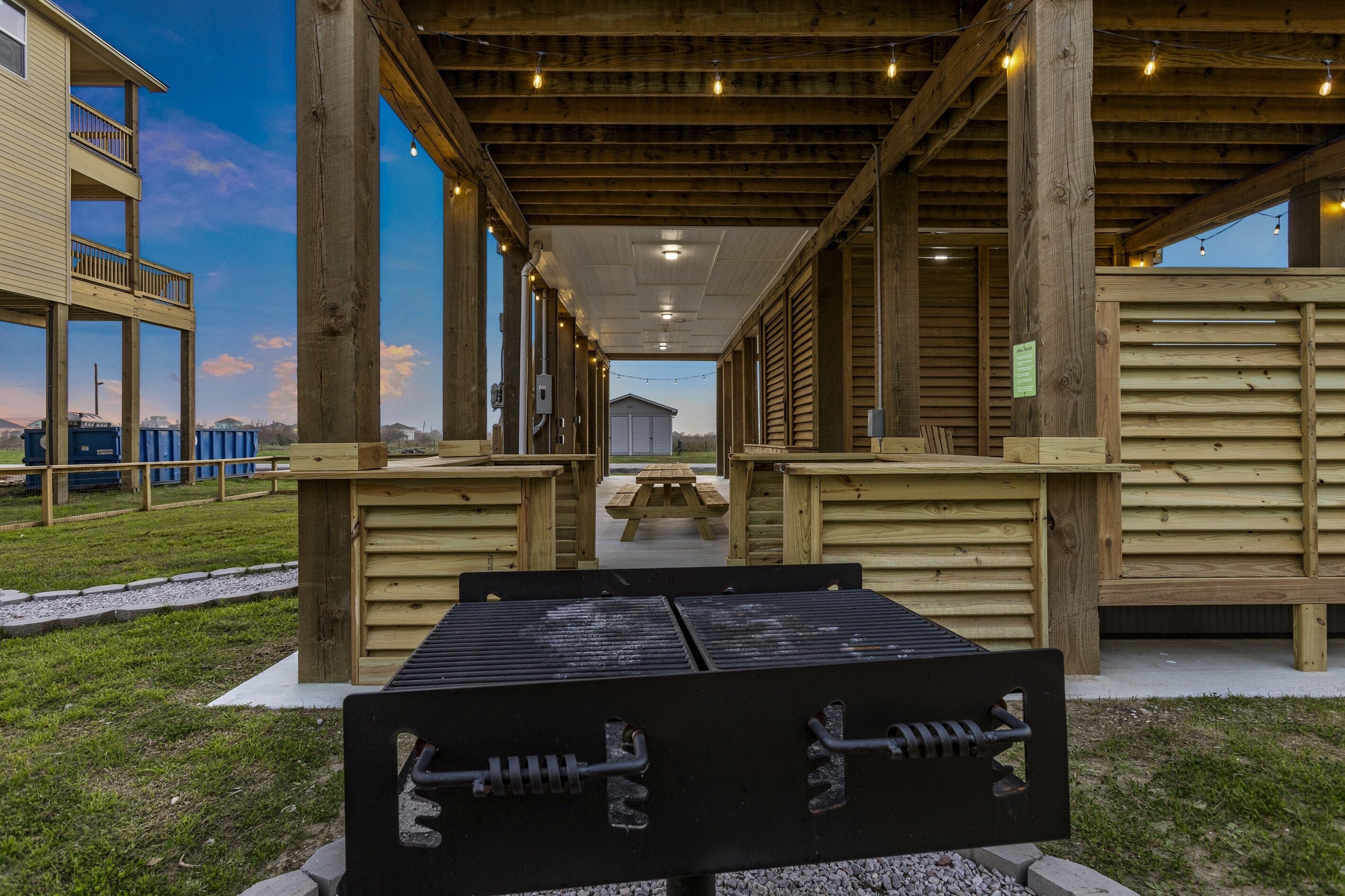 A covered outdoor area with wooden benches and picnic tables, a grill, and a view of a building in the background.