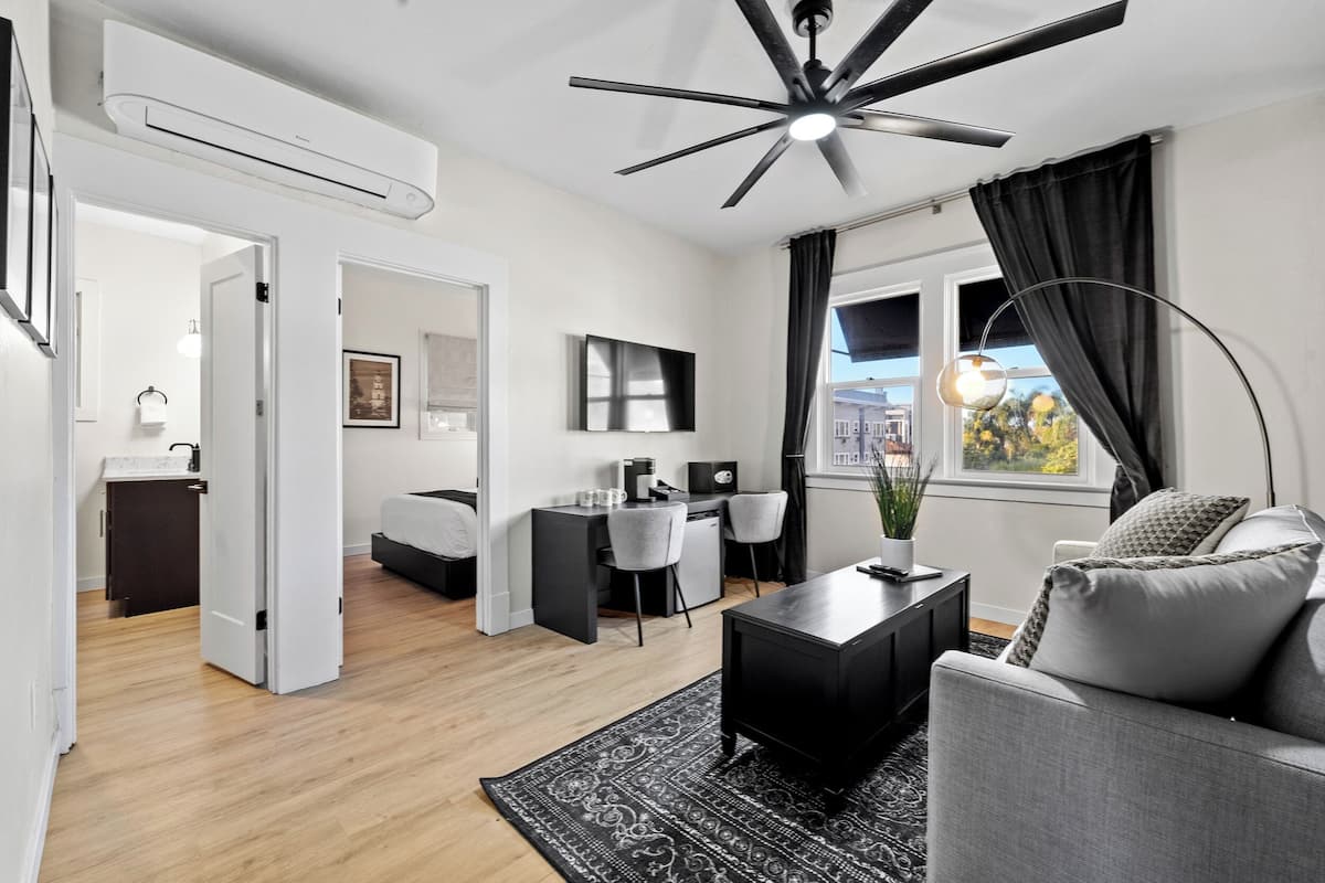 A modern living room with a ceiling fan, a sofa, a coffee table, and a TV. There is a dining area with a table and chairs, and a kitchen in the background.