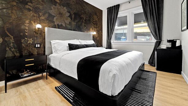 A modern bedroom with a large bed, a black nightstand with drawers, a wall-mounted lamp, and a window with curtains.