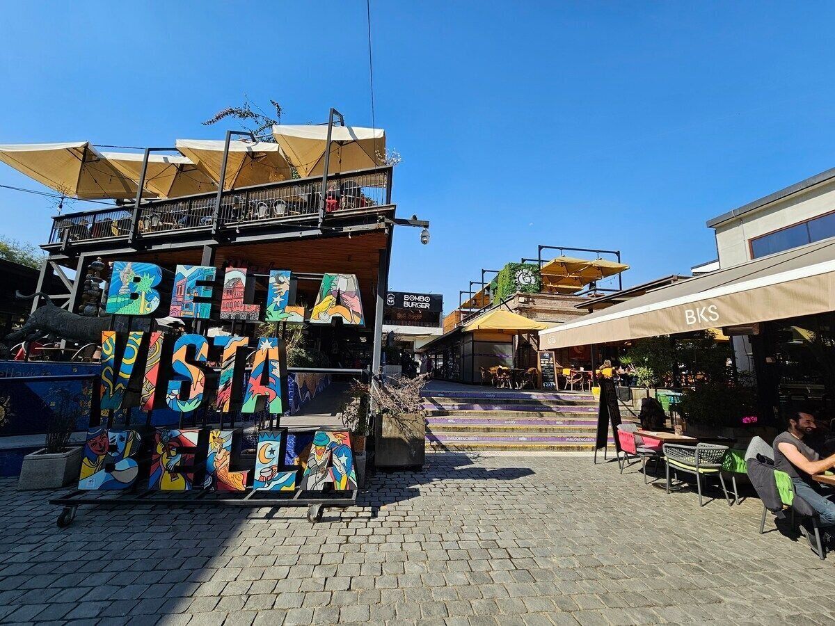 Un espacio exterior con mesas y sillas, un cartel con 'BKS' y obras de arte coloridas sobre un soporte.