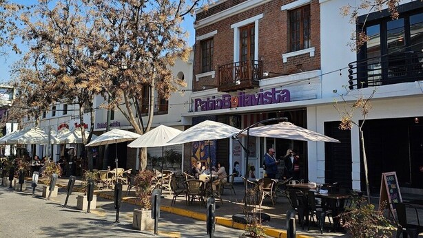 An outdoor café with tables and umbrellas, a brick building with a sign reading "PatioBavisti", and a clear blue sky. - Dardigniac (Santiago)