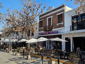 An outdoor café with tables and umbrellas, a brick building with a sign reading "PatioBavisti", and a clear blue sky. - Dardigniac (Santiago)