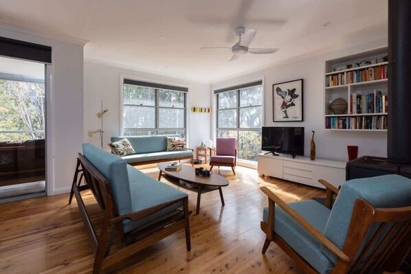 A spacious living room with wooden flooring, a ceiling fan, a sofa, a coffee table, and a bookshelf.