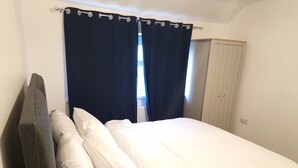 Una cama individual con ropa blanca, una cortina oscura y un armario en una habitación con paredes blancas.