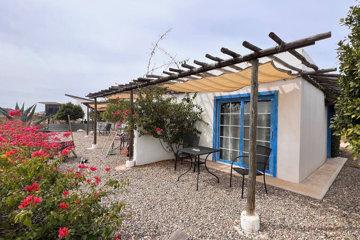 Een klein, laag gebouw met een deur met blauwe kozijnen en een pergola met houten spijlen.