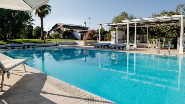 Una piscina cristalina con sillones de descanso y una pérgola, rodeada de follaje y un edificio al fondo.
