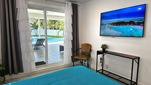 Una habitación con una cama grande, una silla, una mesita pequeña y un televisor mostrando una escena de playa.