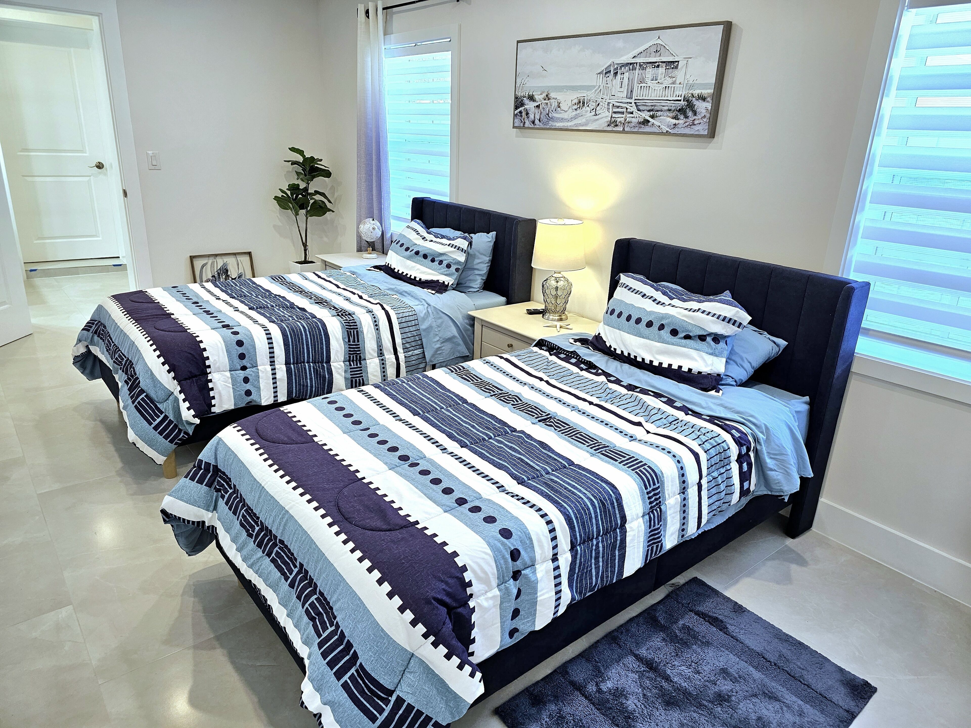 Une chambre avec deux lits, une table de chevet, une lampe, un tableau encadré au mur et une fenêtre avec des stores.