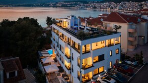 A modern multi-story hotel with balconies, outdoor seating, and a view of a lake and cityscape at dusk. - Beach Villa Valentina Apartment III (Okrug Gornji)