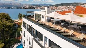 Um edifício moderno com área de piscina no terraço, incluindo cadeiras de descanso e guarda-sóis, com vista para uma cidade costeira e um corpo de água.