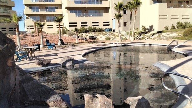 Zona de piscina con una fuente de piedra, sillas de descanso y vistas a un edificio de varios pisos con balcones.