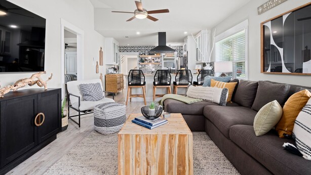 Un salon moderne avec un canapé foncé, une table basse en bois et des chaises noires. On peut voir un ventilateur de plafond et un grand miroir.