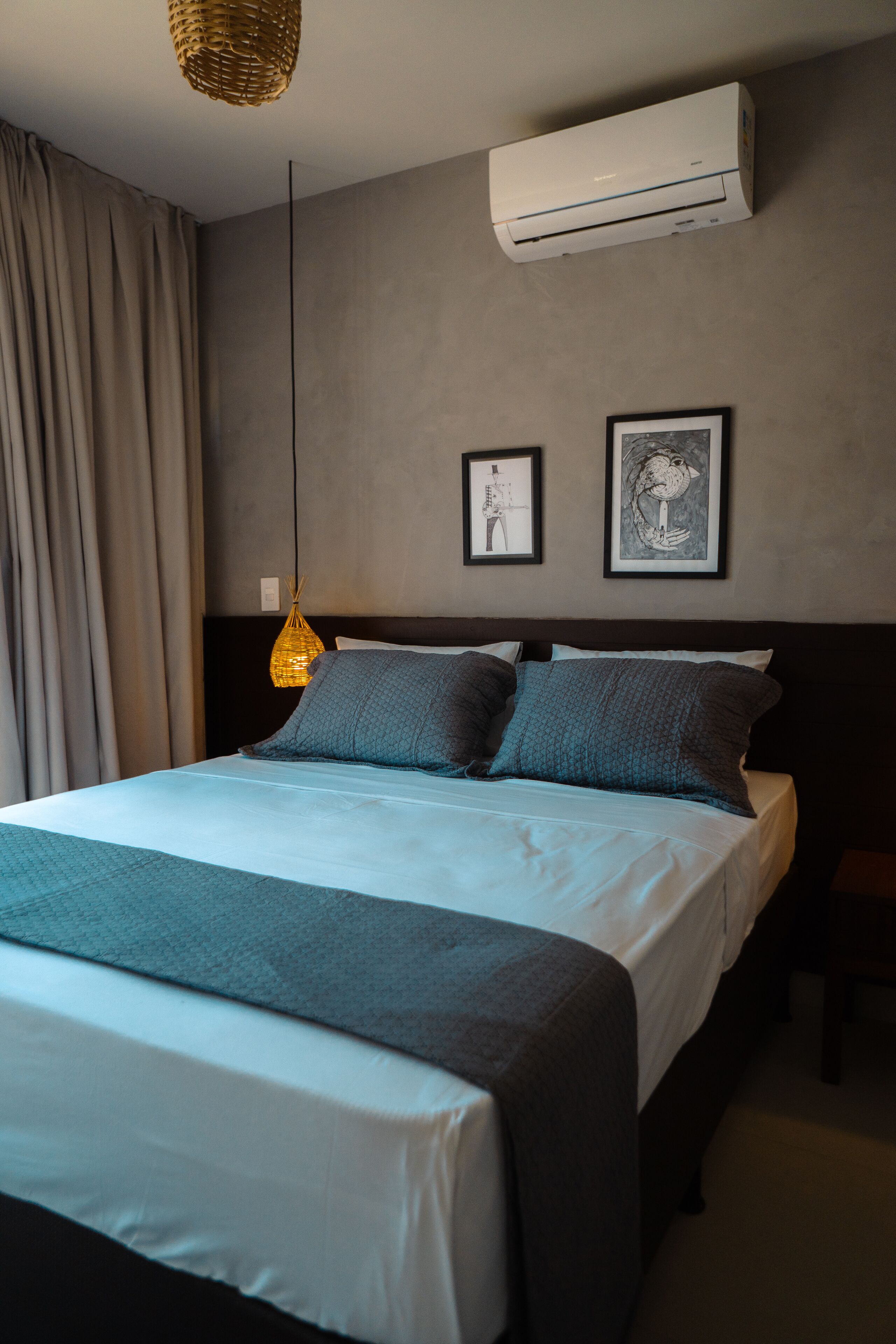 Una cama bien hecha con edredón gris y blanco, una lámpara colgante y dos cuadros en la pared.