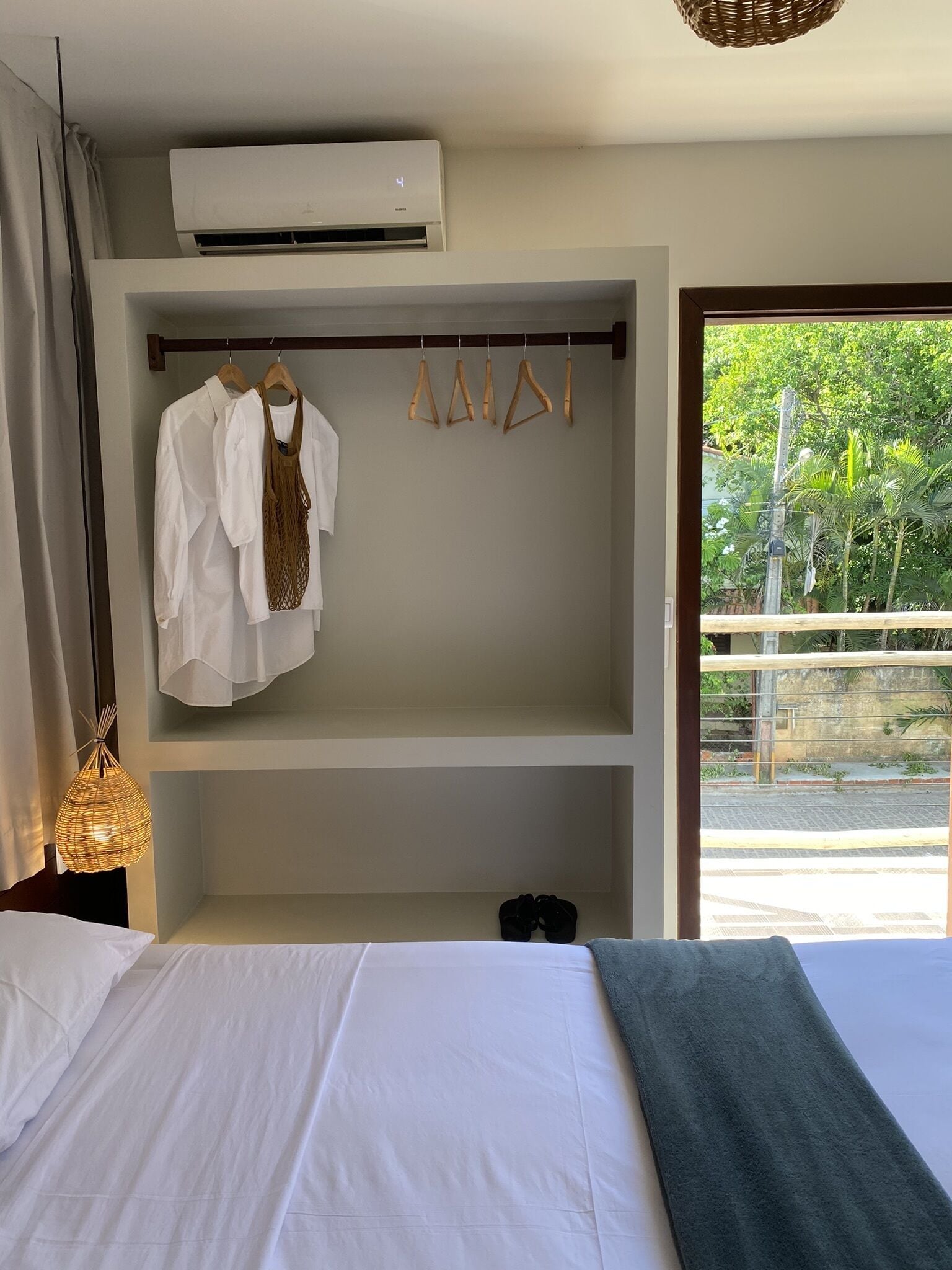 A bedroom with a bed, hanging clothes, an air conditioner, and a view of greenery outside.