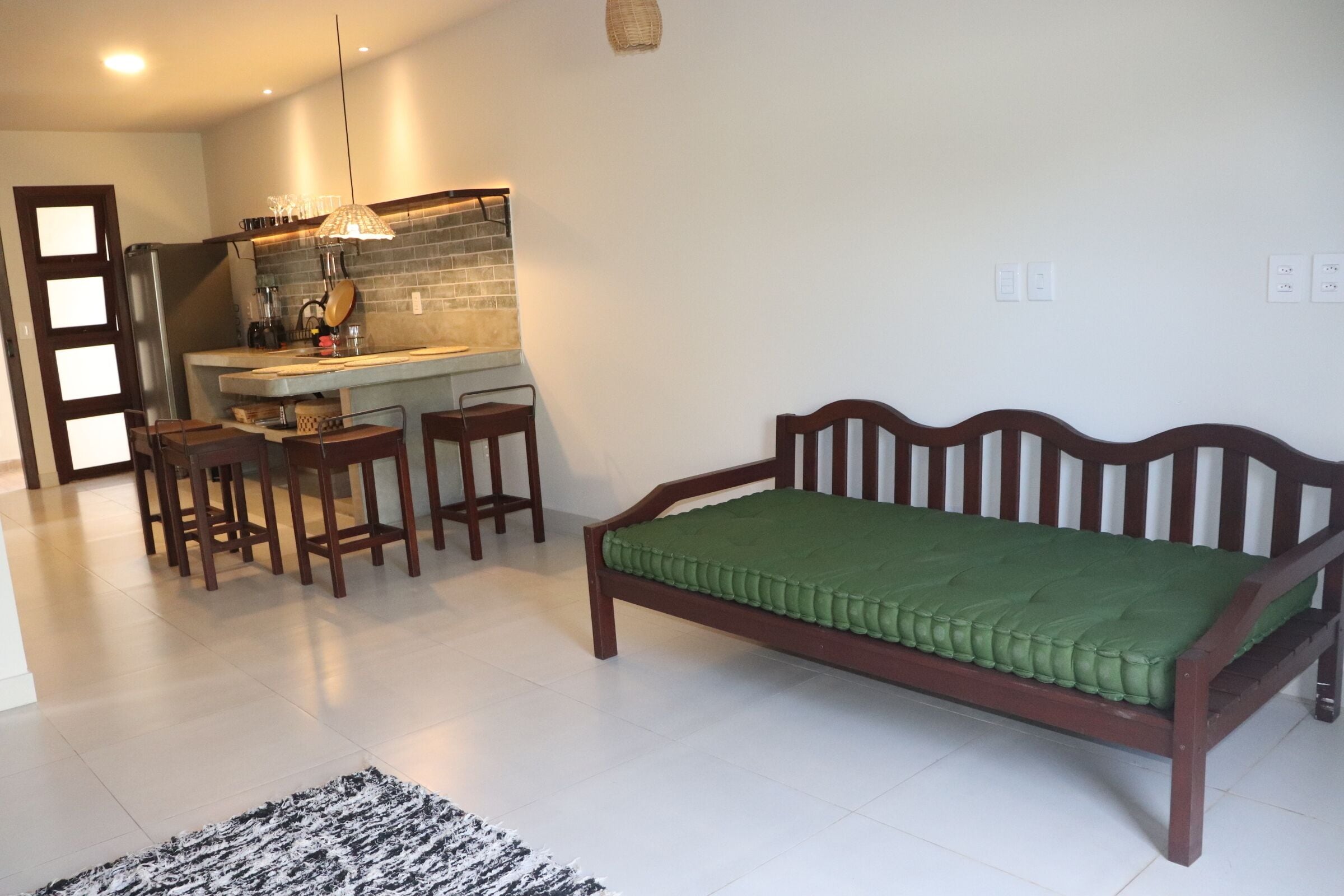 Una habitación con cama de madera, zona de comedor con sillas, cocina con mostrador e iluminación colgante, y suelo de baldosas.