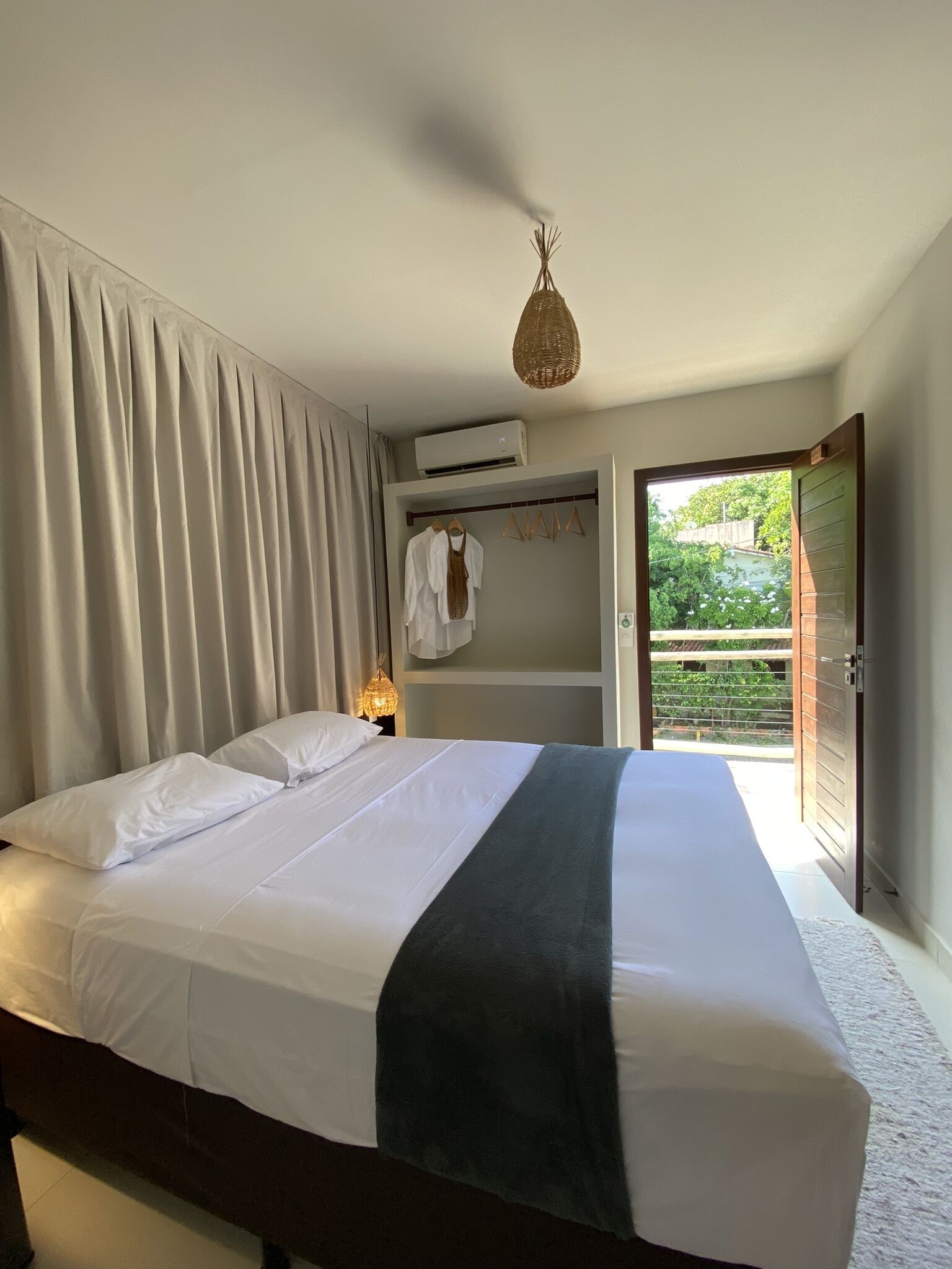 A hotel room with a bed, a hanging light fixture, an air conditioning unit, and a view of greenery outside.
