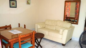 A room with a beige sofa, a wooden dining table with chairs, a small stool, a mirror, and a fan. - Ocean View Apartment B with AC (Bridgetown)
