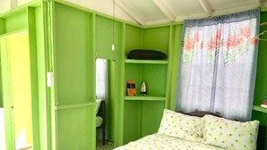 A room with a bed, a shelf with items, and a curtain. - Dover Beach Tiny House w kitchen (Oistins)