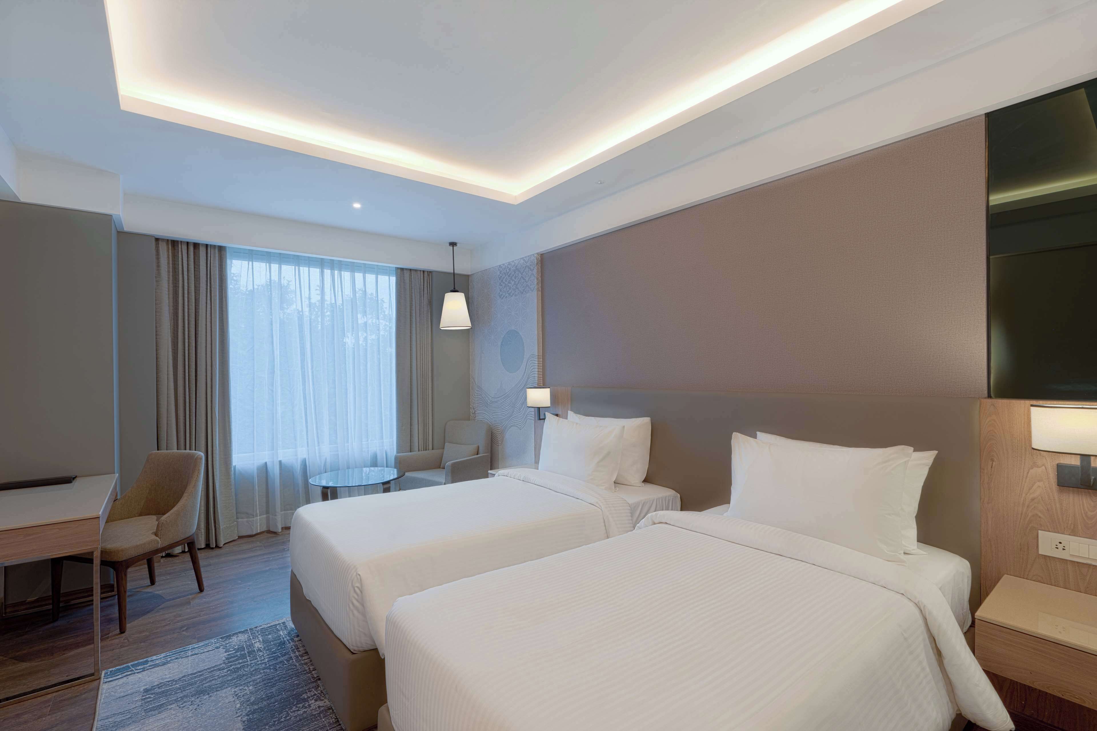 A hotel room with two beds, a desk, a chair, a small table, and a lamp.