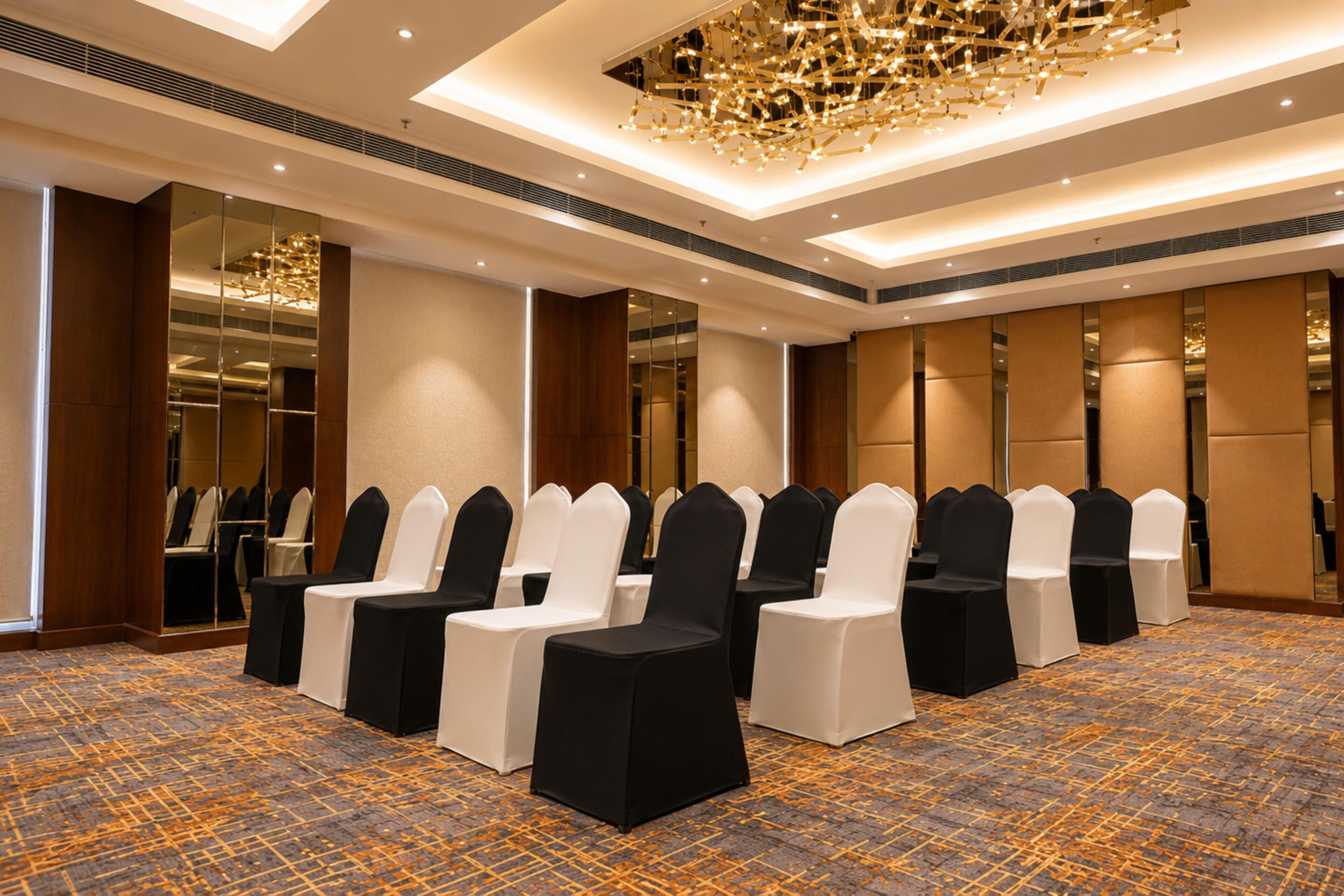 A hotel room with a patterned carpet, a chandelier, and rows of chairs with white and black covers.
