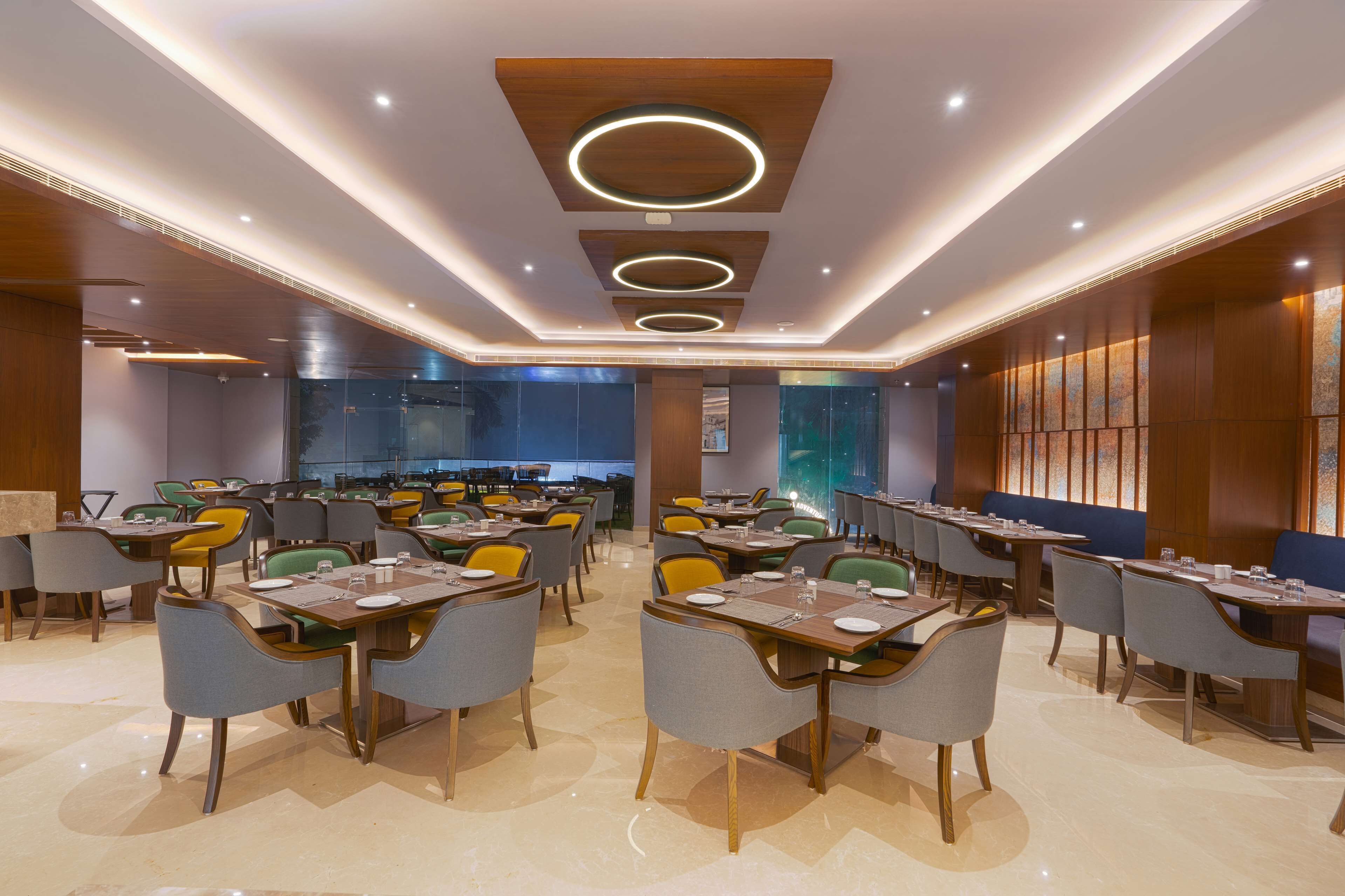 A modern dining area with multiple tables and chairs, a wooden ceiling with circular light fixtures, and a large glass wall in the background.