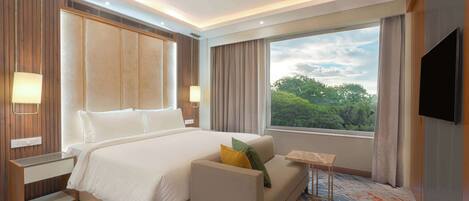 A hotel room with a large bed, a sofa, a TV, and a window with a view of trees.
