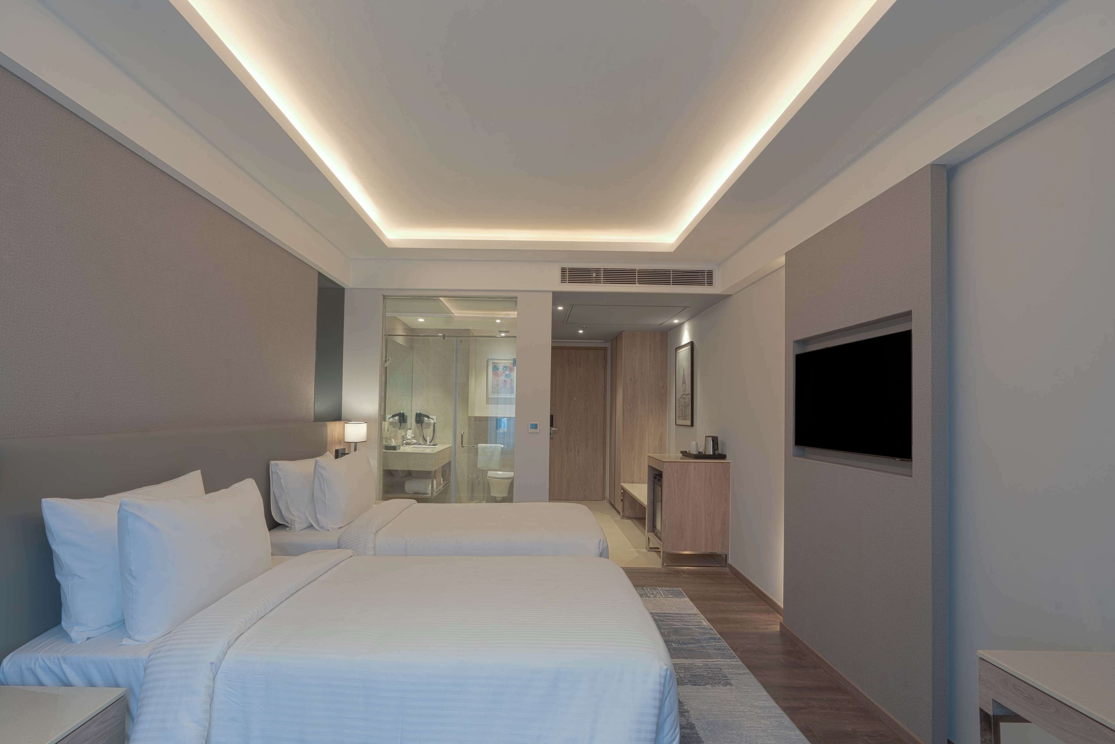 Una habitación de hotel moderna con dos camas, televisión, escritorio y baño visible a través de una puerta de vidrio.
