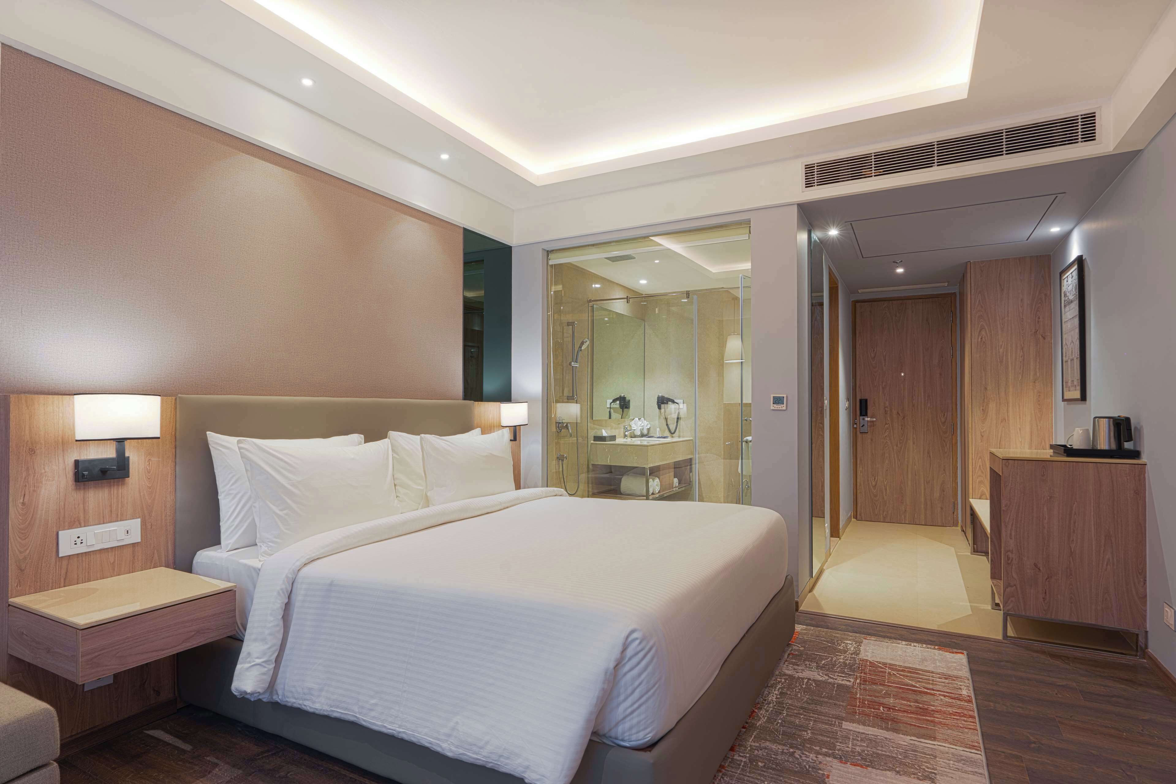 A modern hotel room with a large bed, bedside tables, a built-in wardrobe, and a bathroom with a glass shower enclosure.