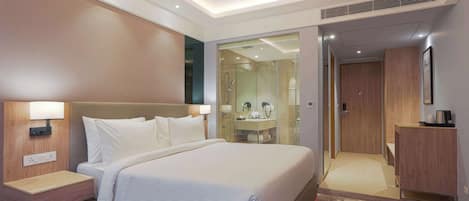 A modern hotel room with a large bed, bedside tables, a built-in wardrobe, and a bathroom with a glass shower enclosure.