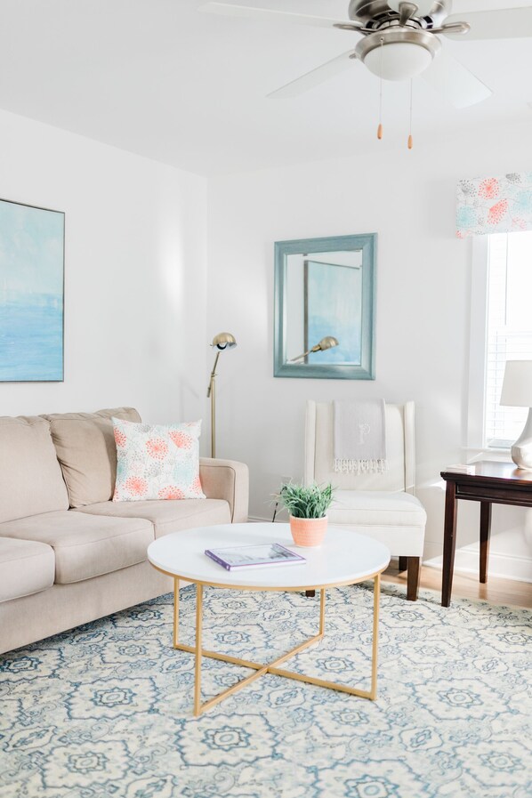 A living room with a beige sofa, a white armchair, a round coffee table, a ceiling fan, and a patterned rug.