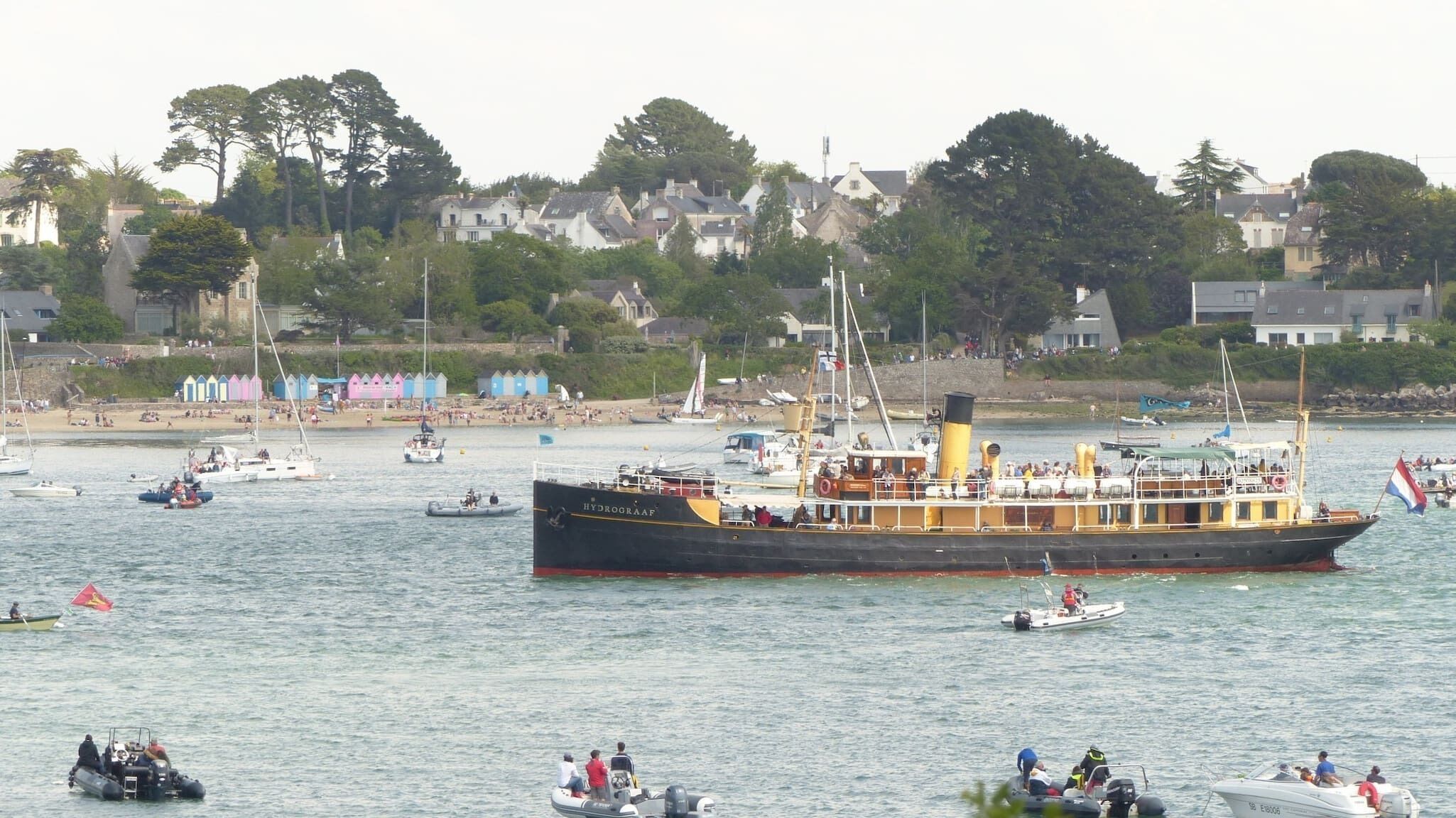 A large ship with multiple decks and passengers, docked at a busy harbor with other boats and a beach in the background.