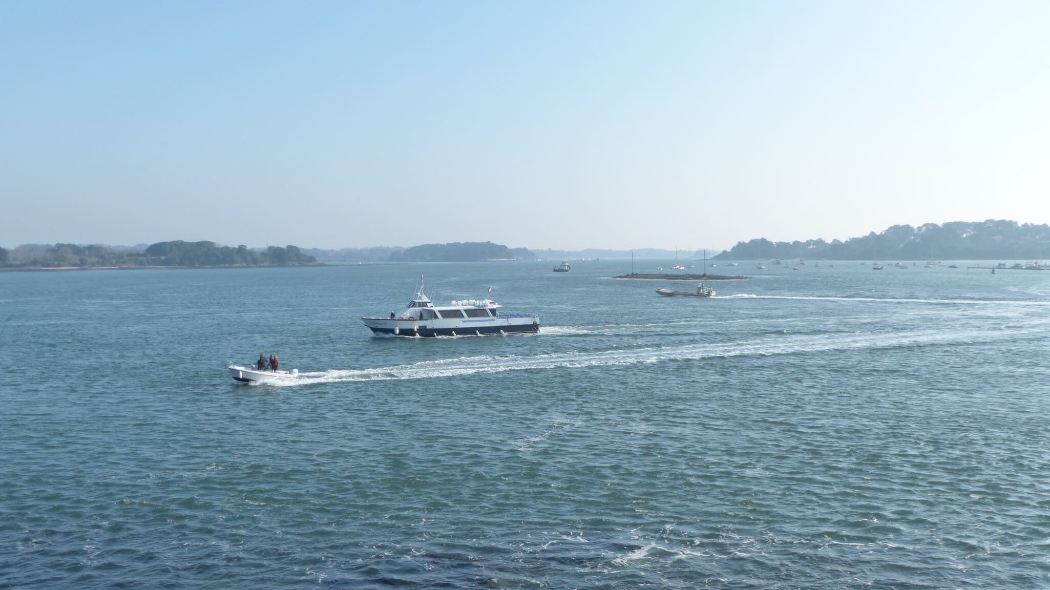 Une étendue d’eau importante où se trouvent plusieurs bateaux, dont un hors-bord et un navire plus grand, ce qui laisse supposer une voie navigable ou une marina très fréquentée.