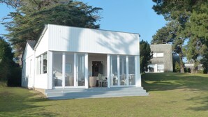 A modern, single-story building with a flat roof and large glass windows. It features a covered porch with a few chairs and a table. - Maritime cottage with exceptional sea view (Baden)