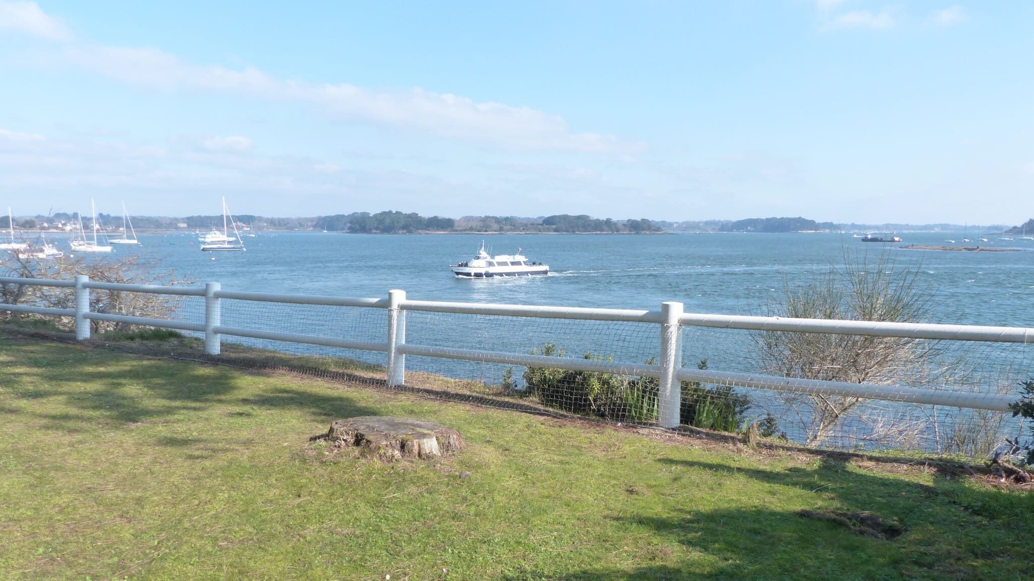 A waterfront view with a white fence, a grassy area, and a body of water with boats.