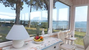A room with a large window offering a view of a body of water and distant mountains. There is a table with a lamp, a bowl of fruit, and a chair. - Maritime cottage with exceptional sea view (Baden)