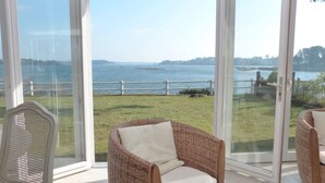A room with a large glass door leading to a grassy area with a view of a body of water and a distant island. - Maritime cottage with exceptional sea view (Baden)