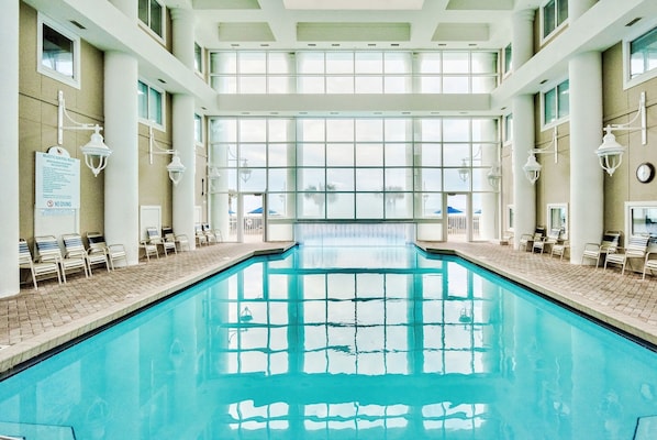 An indoor swimming pool with clear glass walls, multiple windows, and lounge chairs along the sides.
