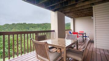 A covered outdoor patio with a wooden table and chairs, a view of a forested area, and a white wall in the background.