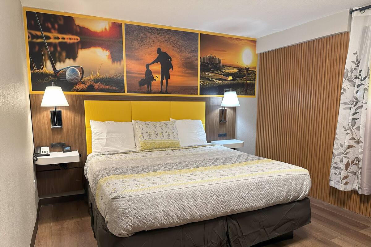 A hotel room with a large bed, two bedside tables, a wooden headboard, and wall art depicting golf and sunset scenes.