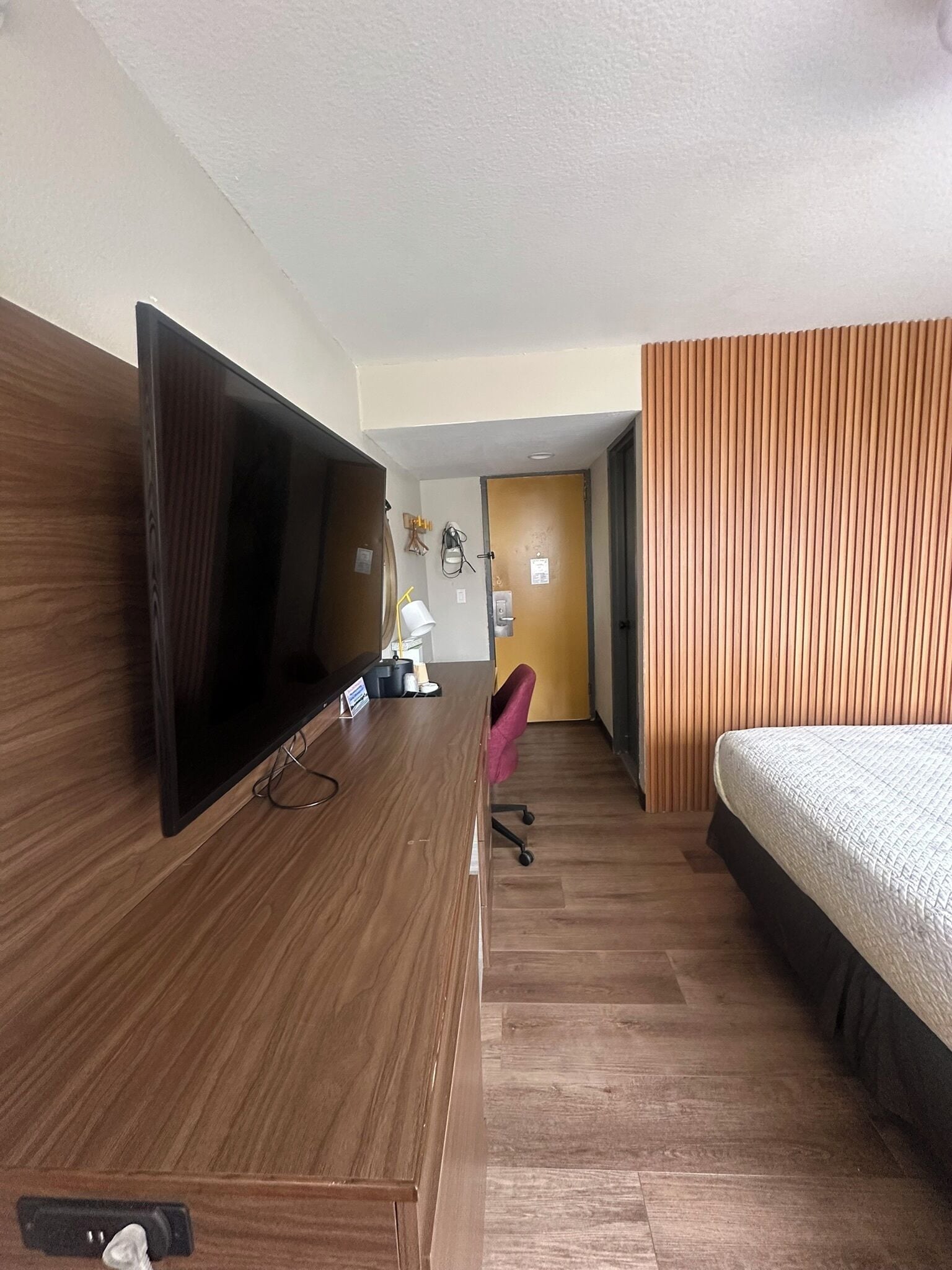 Quarto de hotel com escrivaninha de madeira, televisão de tela plana, cama e banheiro com porta de madeira.