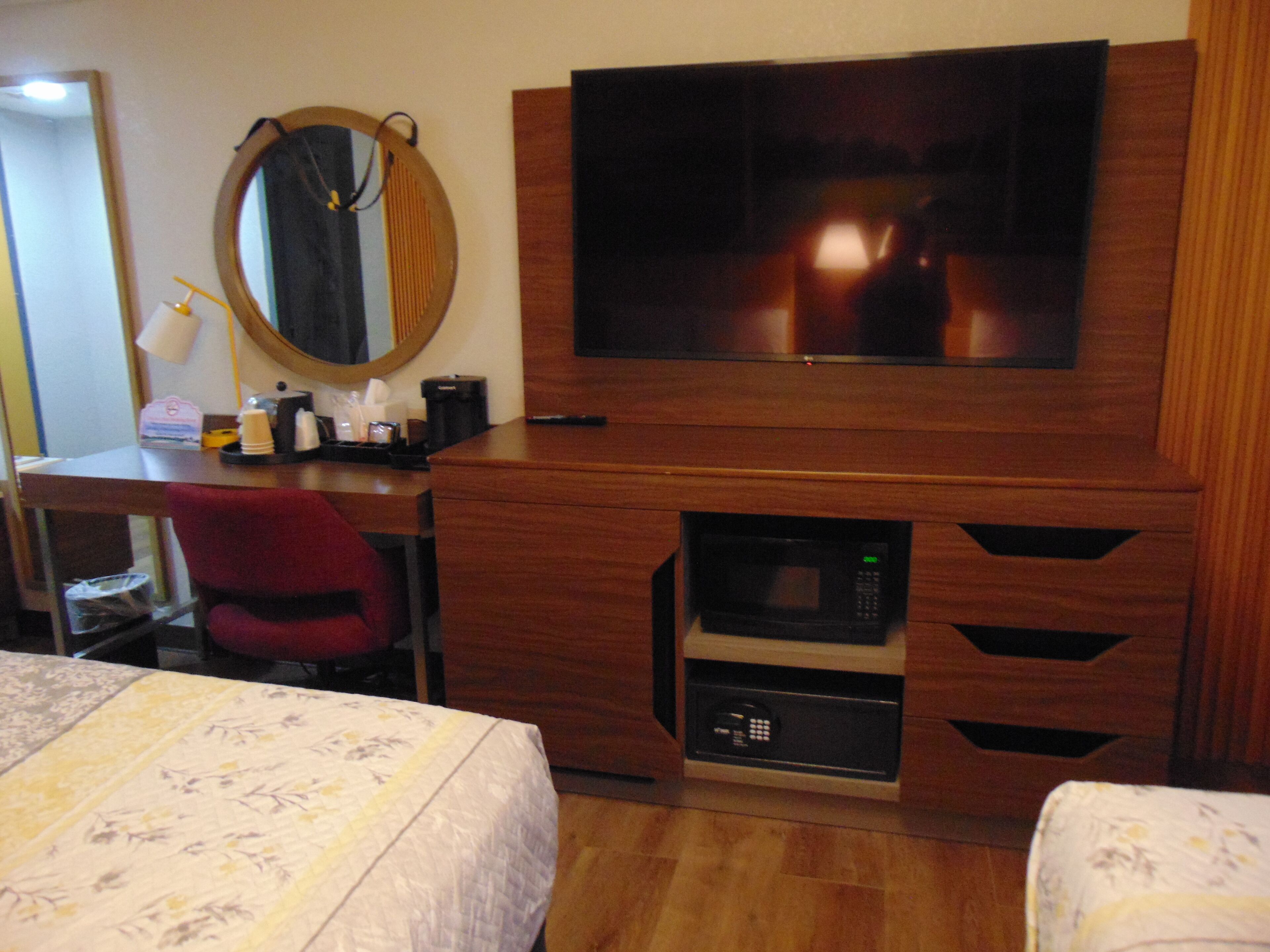 A hotel room with a wooden desk, a television mounted above it, a microwave built into the desk, a red chair, and two beds with floral bedspreads.