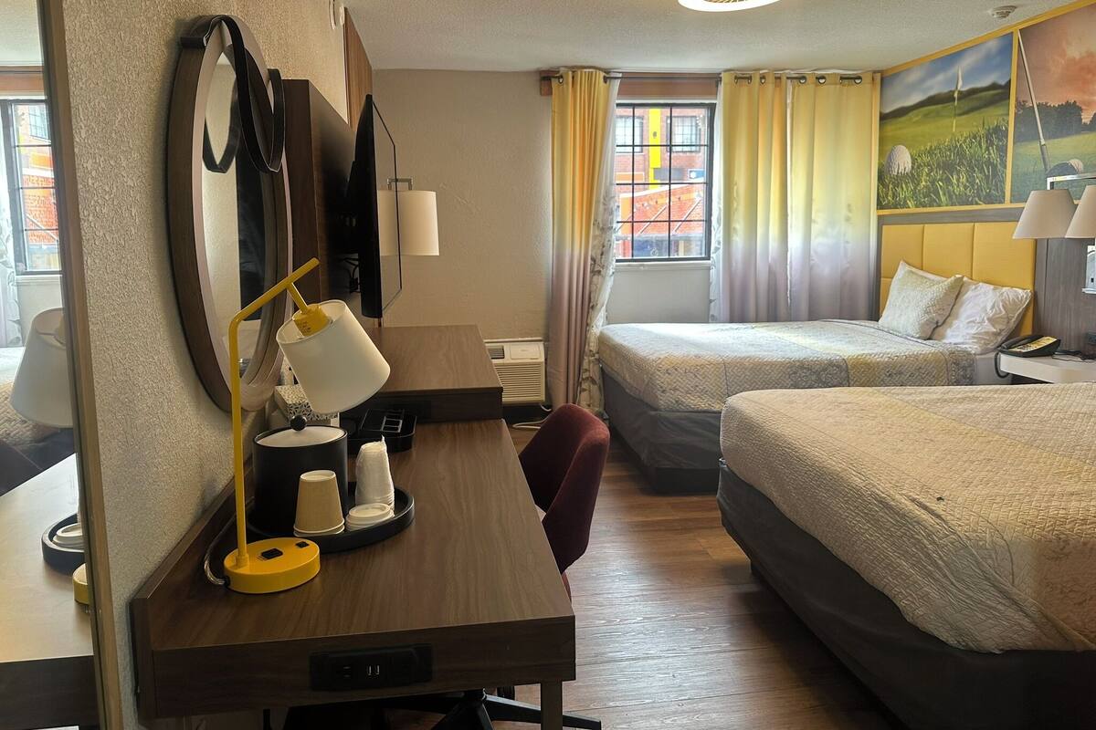 A hotel room with a wooden desk, two beds, a large mirror, and a window with curtains.
