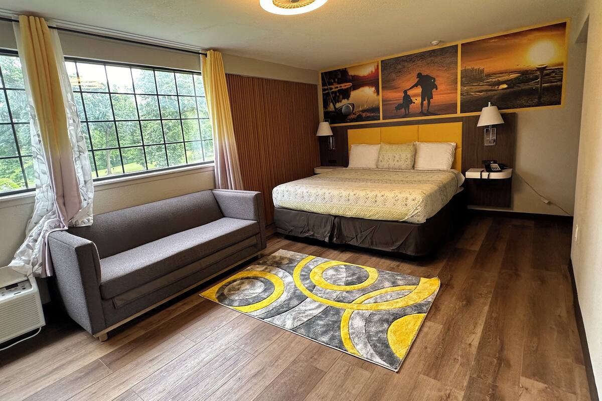 A hotel room with a bed, a sofa, a window with curtains, a wall with artwork, and a floor lamp.