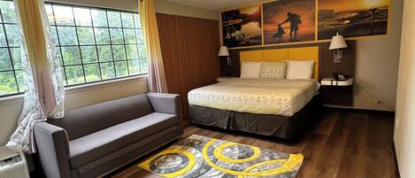 A hotel room with a bed, a sofa, a window with curtains, a wall with artwork, and a floor lamp.