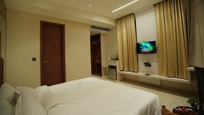 A hotel room with a bed, a TV mounted on the wall, a small bedside table, and a window with curtains. - Blue Diamonds Inn (Greater Noida)