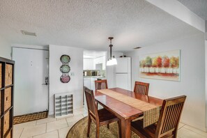 A dining area with a wooden table and chairs, a kitchen in the background, and a painting on the wall. - New Smyrna Beach Condo w/ Ocean-View Balcony! (New Smyrna Beach)